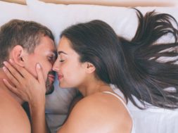 Cheerful and nice couple is lying in bed close to each other. Girl holds her hand on his neck. They are touching their noses. This picture is very nice and tender