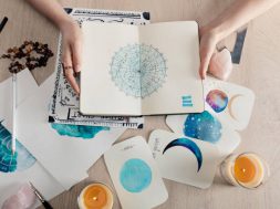 Top view of astrologer holding notebook with watercolor drawings and zodiac signs on cards on table