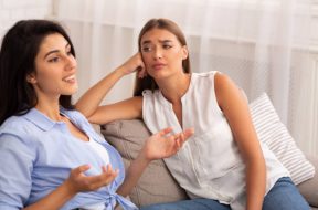 Envy. Jealous Girl Listening To Her Friend Talking About Her Great Life Sitting On Couch At Home. Selective Focus