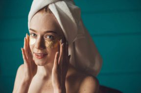 Caucasian freckled woman applying golden collagen patches under her eyes, looking at camera. Natural beauty. Facial treatment.