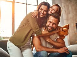 Happy dad and mom with their cute daughter and teddy bear hug and have fun sitting on the sofa in the living room at home.