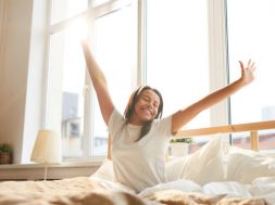 Portrait of beautiful Mixed-Race woman stretching sitting in bed lit by sunlight, copy space